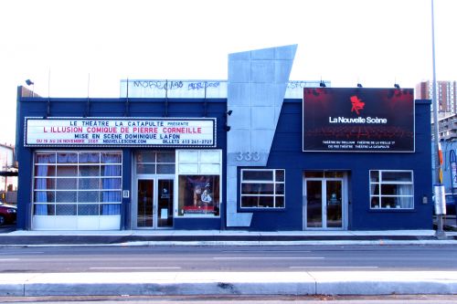Colour photograph of a one-story brick theatre building, painted dark blue, with several glass doors and windows. A marquee announces that the next play will be “L'Illusion comique de Corneille,” presented by the Théâtre la Catapulte.