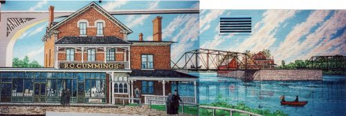Photographie en couleur d’une murale illustrant, de près et de loin, le commerce en brique « R.C. Cummings ». Ce dernier se situe sur une île, au centre d’une rivière traversée par un pont en métal.