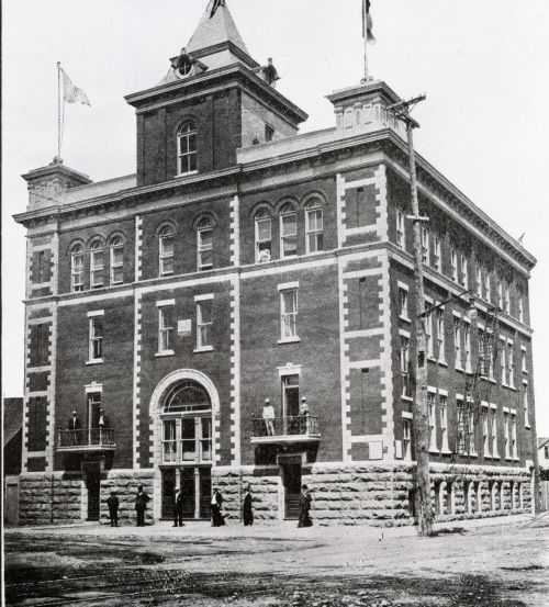 Black and white photograph of a four-storey brick building. The entrance door is topped by a tower, and two turrets are positioned at the front corners of the building. Flags float at the top of the turrets. A few men stand in front of the building; others stand on the first-floor balconies.