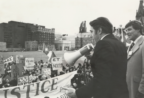 Black and white photograph of two middle-aged men. One is holding a megaphone. They are speaking to a crowd gathered for a manifestation, on Parliament Hill.