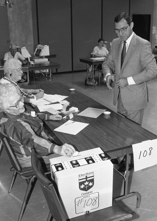Black and white photograph of a mature, dark-haired man wearing glasses, and a suit and tie. He is standing in front of a polling station run by two elderly women. One of them is inserting a ballot into the ballot box. In the background, other polling stations.