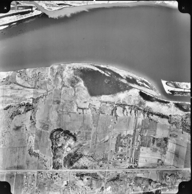 Black and white aerial photograph of a rural landscape with a river to the north (at the top of the image). A few dwellings are grouped to the south, along two main roads (at the bottom of the image).