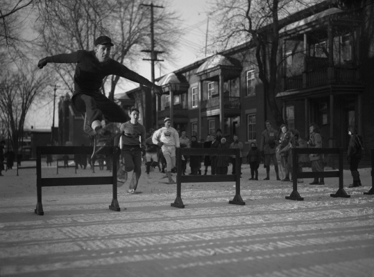 Photographie en noir et blanc de trois jeunes hommes qui participent à une course à obstacles en raquettes, dans un parc urbain. À l’avant-plan, un jeune homme en plein saut. Des spectateurs regardent la course.