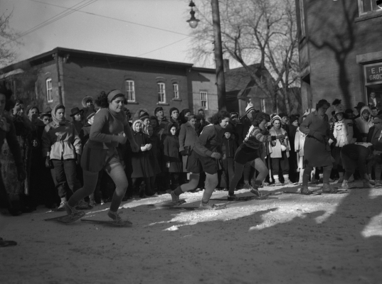 Photographie en noir et blanc de trois jeunes femmes et d’une femme d’âge moyen qui participent à une course en raquettes, en pleine ville. Derrière elles, plusieurs personnes regardent la course. À l’arrière-plan, on voit des maisons.