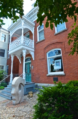 Colour photograph of a two-storey red brick building. The front is decorated with a white-washed wood balcony. In front of the entrance, a sculpture of a stylized head in profile. In the window, a table and the name of the business, “GJCB Galerie Jean-Claude Bergeron.”