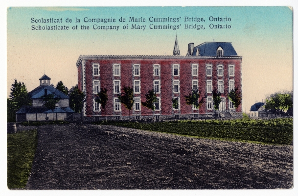 Colour postcard with text printed in French and English. A dirt road leads to a three-storey red brick building topped by a bell tower. Small trees grow alongside the building. On one side of the structure, an outbuilding; on the other side, a small house.