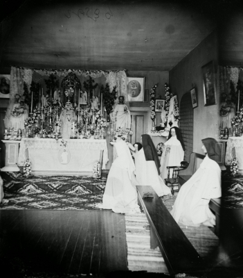 Black and white photograph of a novice and four nuns praying in a modest chapel. The altar is decorated with candlesticks, flowers, statues and holy images.