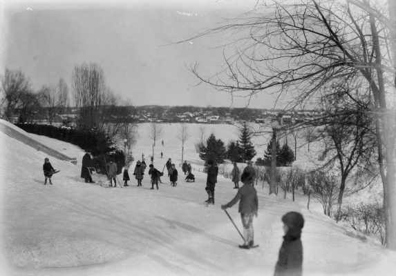 Photographie en noir et blanc d’un paysage hiver où se retrouvent jeunes et moins jeunes. Certains jouent au hockey et d’autres montent et descendent une côte avec des traîneaux. À l’arrière-plan, une rivière.