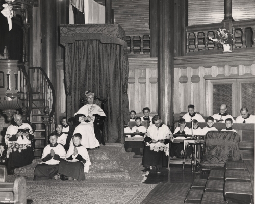 Black and white photograph of a mature cleric wearing the biretta, sitting in a raised chair next to the pulpit. He is surrounded by five other clerics and young altar boys, all dressed in cassocks and surplices.