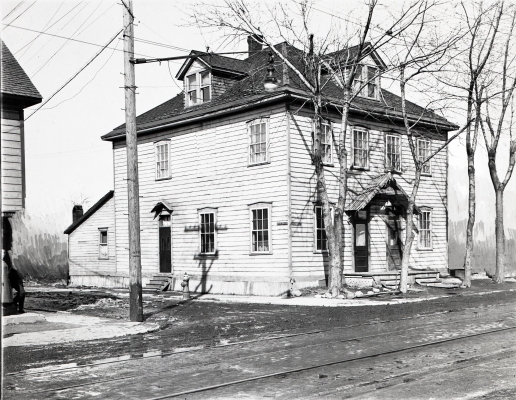 Black and white photograph of a two-storey wooden house, viewed from the side. Two dormers are set into the roof. Bare trees grow along the front of the house. There is snow on the ground.