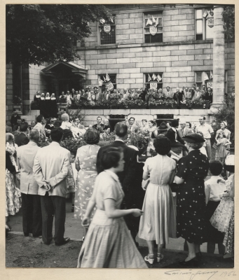 Photographie en noir et blanc d’un attroupement devant un édifice en pierre, aux fenêtres richement décorées. Le groupe inclut des hommes et des femmes ainsi que des religieuses et des enfants.