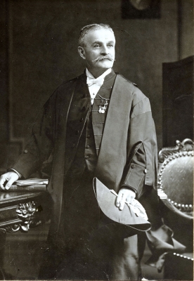 Black and white photograph of an elderly man standing in an office with rich furniture. The man wears the parliamentary attire of Speaker of the Senate, as well as military medals.