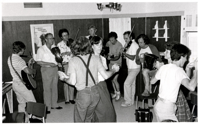 Photographie en noir et blanc d’une soirée dansante. Deux couples de jeunes adultes dansent au centre d’un demi-cercle formé par quatre violoneux, deux guitaristes et un accordéoniste. Les musiciens sont tous des hommes.