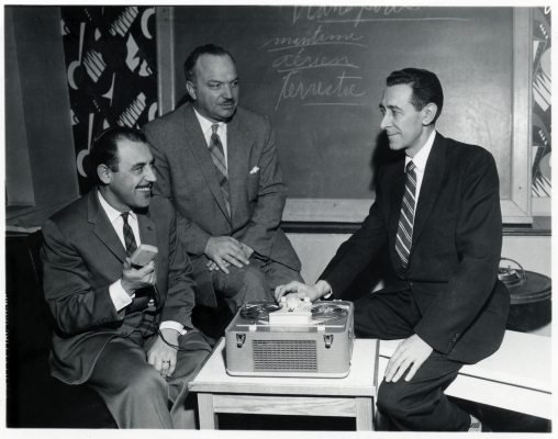Photographie en noir et blanc de trois hommes d’âge mûr en costumes et cravates. Ils sont assis autour d’un magnétophone à bobines. L’un deux arbore un grand sourire. Derrière eux, un tableau noir.