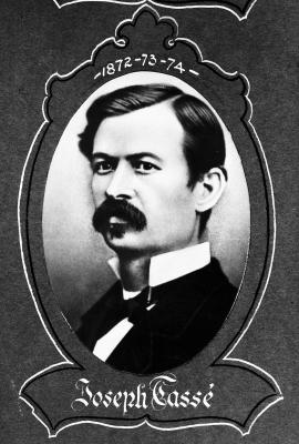 Black and white studio photograph of a young man with a mustache and a severe look. The man, appearing in three-quarter view, wears a black jacket and a bow tie. The photograph, oval in shape, is surrounded by a decorative frame. Name and dates are indicated.