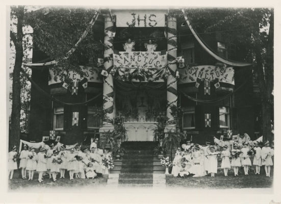 Black and white photograph of an altar at the front of a three-storey house, decorated with banners and streamers. About thirty children – dressed in white, wearing crowns and wings, holding bouquets of flowers – stand or sit on either side of the stairway leading to the altar.