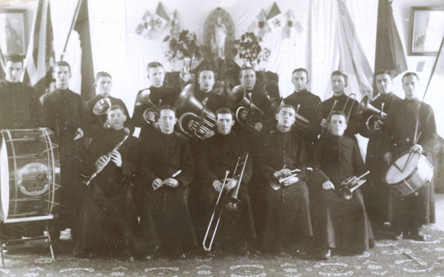 Photographie en noir et blanc d’un groupe d’une quinzaine de musiciens, d’âges divers. Cinq d’entre eux sont assis, les autres sont debout derrière. Tous portent la soutane et tiennent leur instrument.
