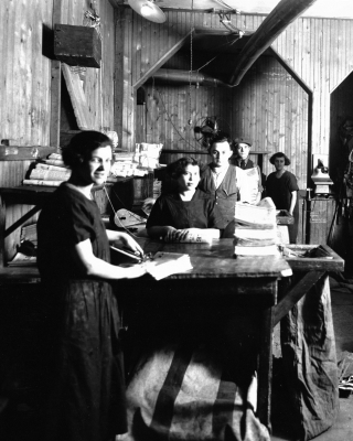 Black and white photograph of three young women and two young men standing at counters. They are surrounded by stacks of newspapers.