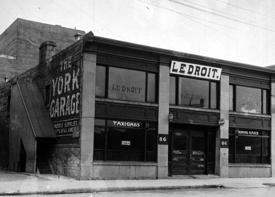 Black and white photograph of a two-storey brick building with many windows. The sign “Le Droit” is hung on the second floor. The words “The York Garage” appear on the side of the building. Signs over the first-floor windows read “TAXI CABS” and “TOURING CARS.”