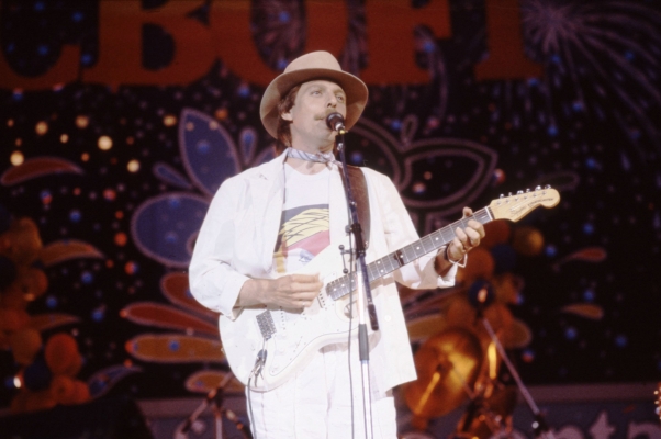 Colour photograph of a middle-aged man with a mustache on a stage. He wears a hat, a scarf around his neck, and a white suit. He plays the electric guitar and sings in front of a microphone. In the background, geometric patterns on colourful background and the words “CBOF-FM.”
