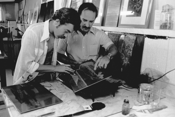 Black and white photograph of a middle-aged man and a younger man in an art studio. The two men are examining a work of art. Several paintings are arranged on the floor and the walls.
