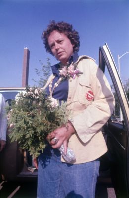 Colour photograph of a young woman in casual dress, wearing a corsage and a red button on her jacket. She is getting out of a car, a bouquet in her hands.