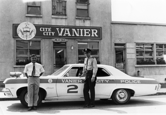 Black and white photograph showing a smiling man and a serious-looking policeman by a police car in the town of Vanier. The car is parked in front of a Vanier building.