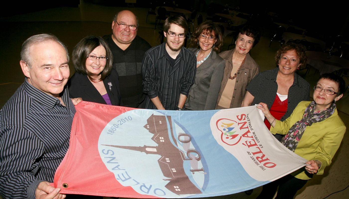 Colour photograph of a group of people, including two older man, two older women, three middle-aged women, and a young man. The group is gathered around a colourful banner commemorating the 150th anniversary of Orléans.