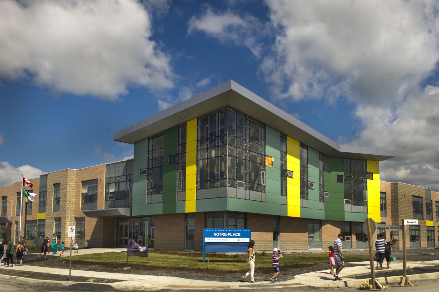 Colour photograph of a two-storey brick and glass building with coloured sections surrounding the glass. Children and parents walk around the building. In the centre, a blue and white sign bearing the name of the school.