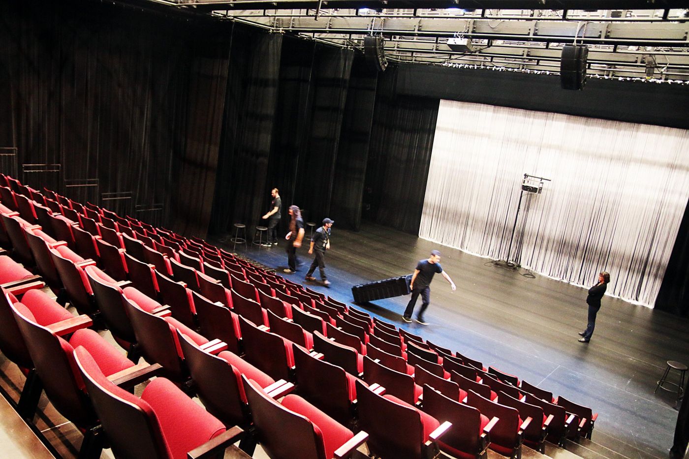 Photographie en couleur, prise de biais. Une salle de théâtre vue d’en haut, à partir des rangées de sièges rouge vif disposées en gradins. Cinq techniciens vêtus en noir travaillent sur la scène.
