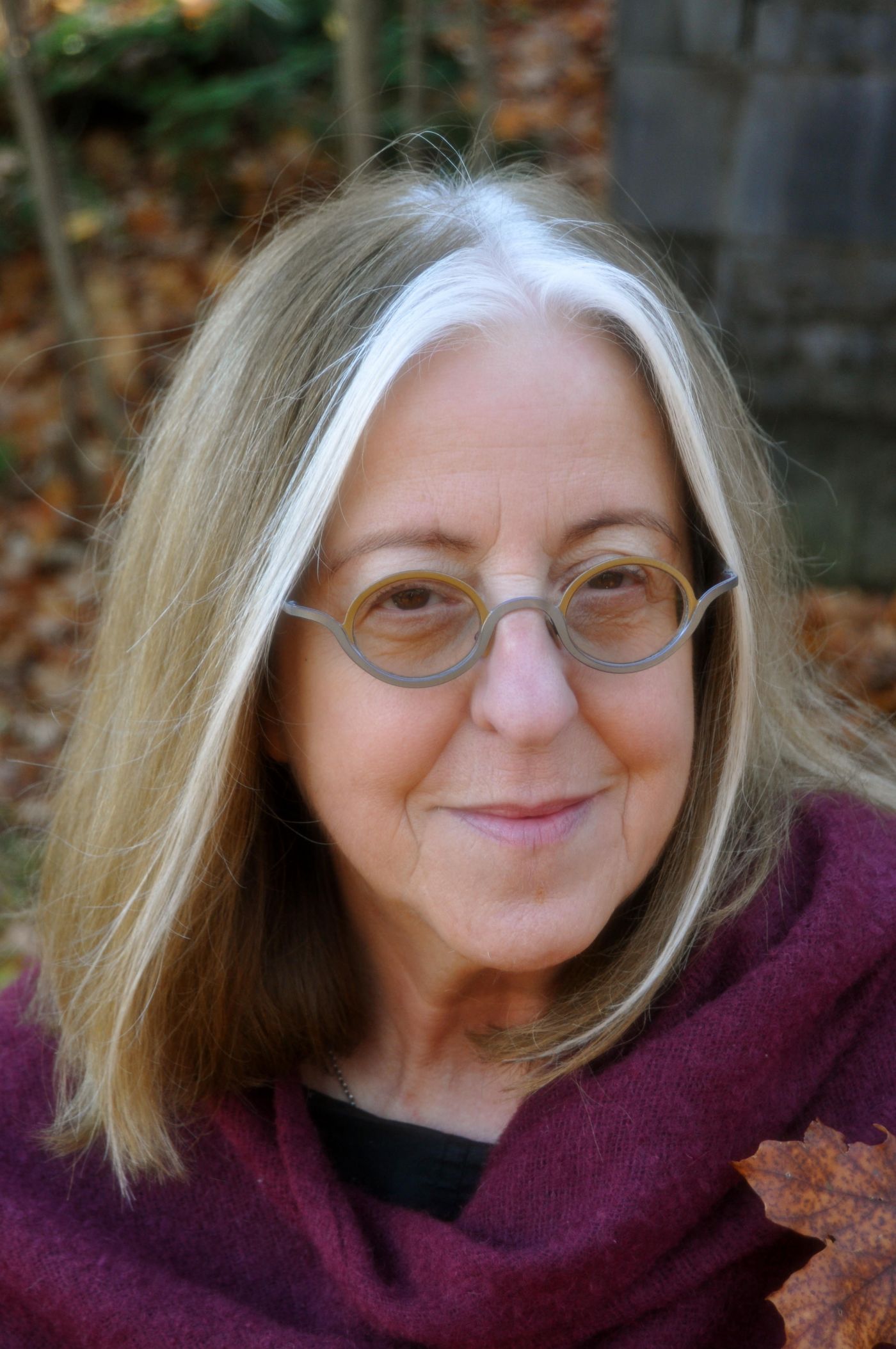 Close-up colour photograph of an older woman with glasses and medium-length salt-and-pepper hair. She wears a purple scarf in the outdoor autumn setting. She smiles at the camera.