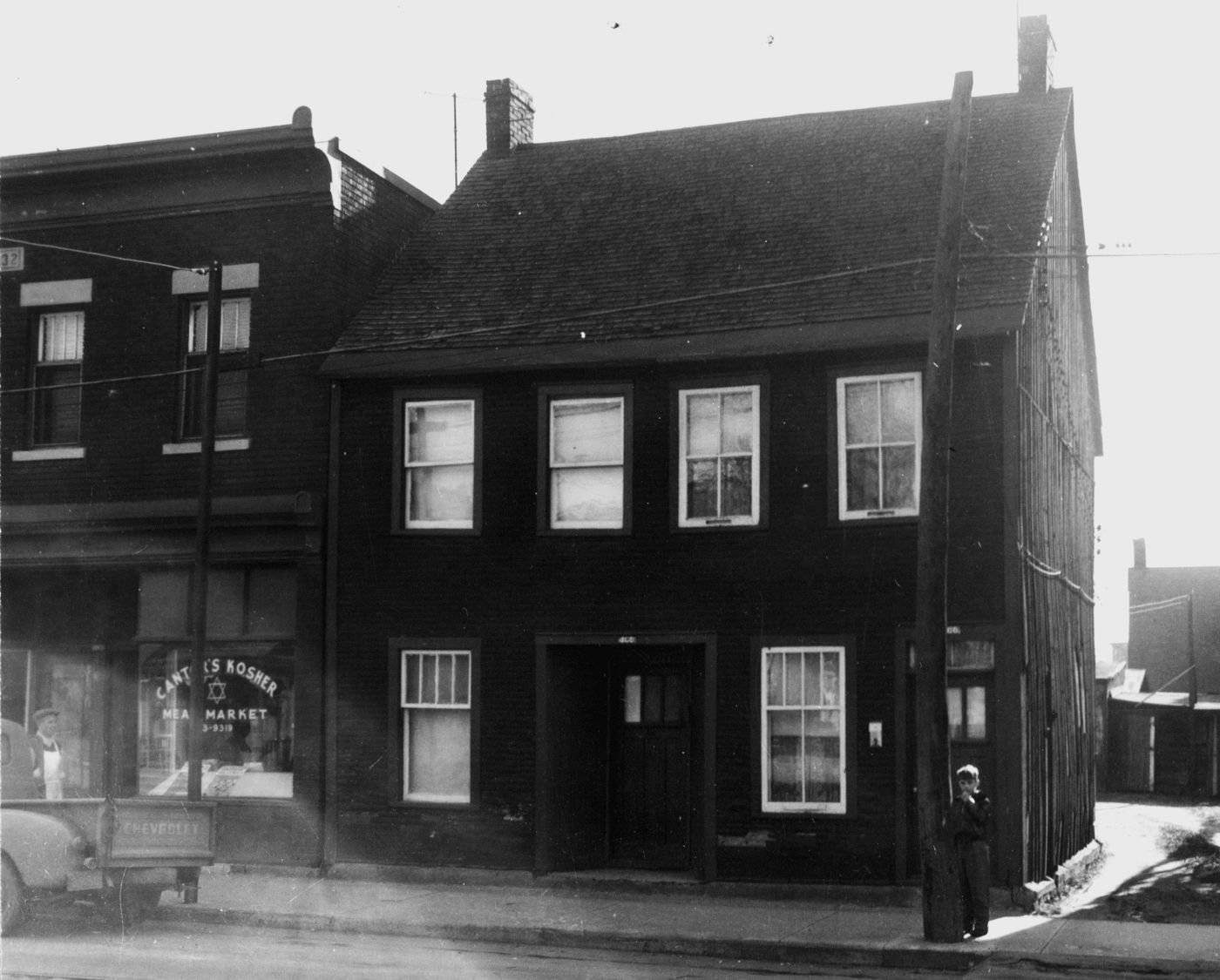 Photographie en noir et blanc d’un jeune garçon devant une maison en bois à deux étages. Un camion et un homme sont devant le commerce Cantor's Kosher Meat Market.
