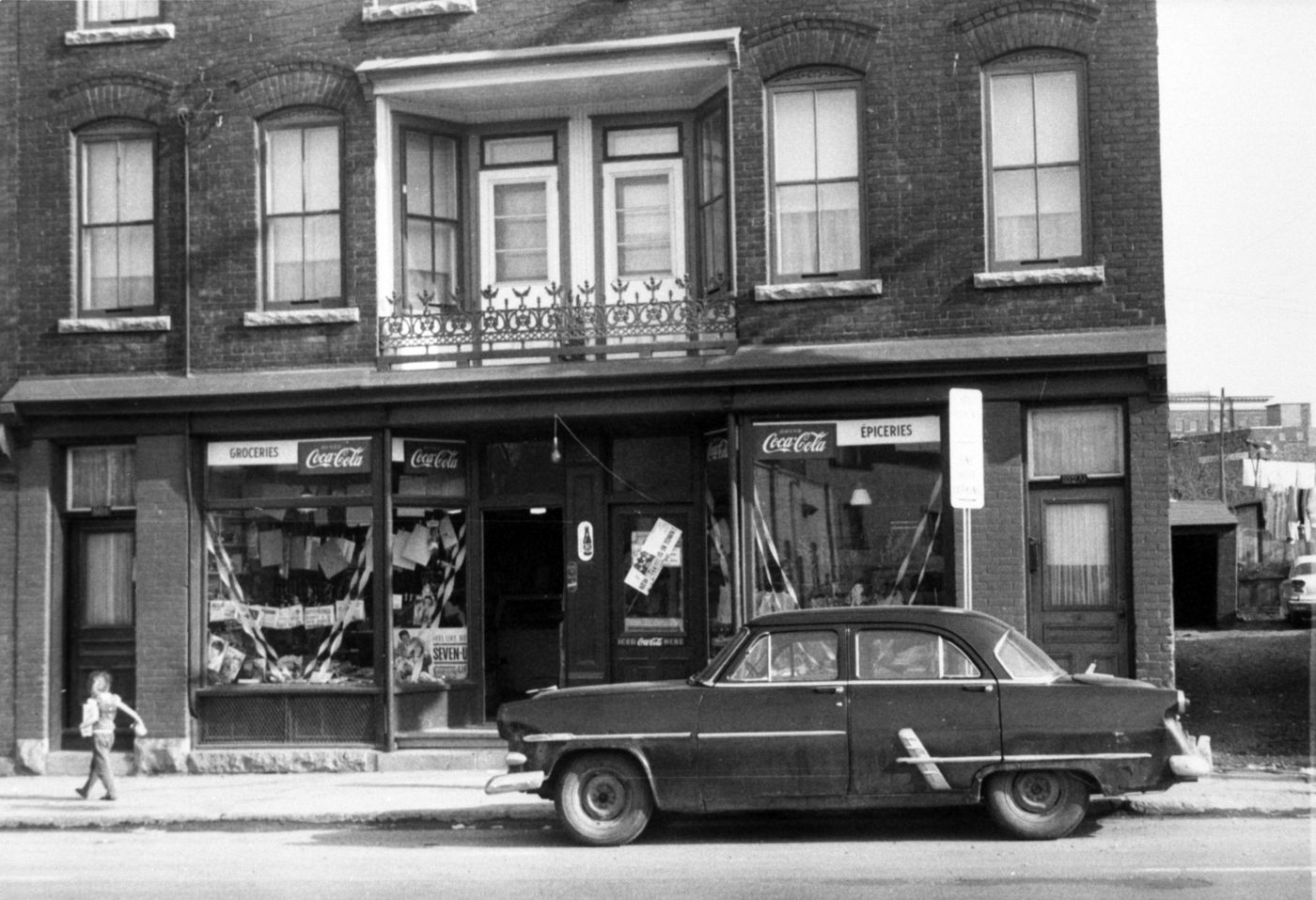 Black and white photograph of a young boy walking in front of a multi-storey brick house, with a grocery store on the ground floor. The signs are bilingual. A car is parked in front of the shop.