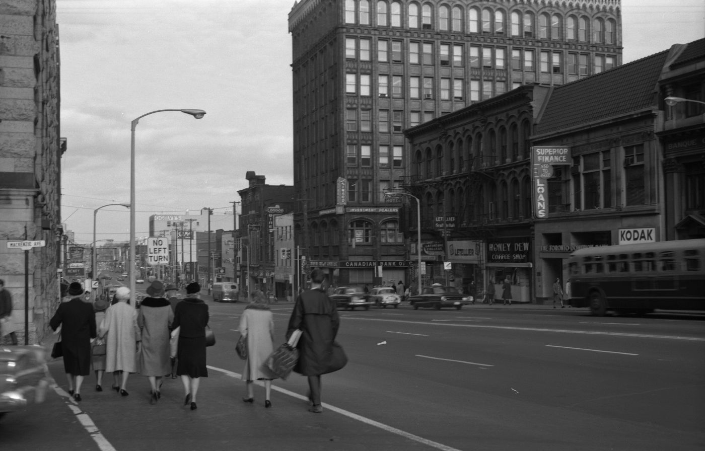 Photographie en noir et blanc d’une rue passante avec édifices de plusieurs étages et panneaux publicitaires. À l’avant-plan, un groupe de piétons bien mis traverse une rue transversale.
