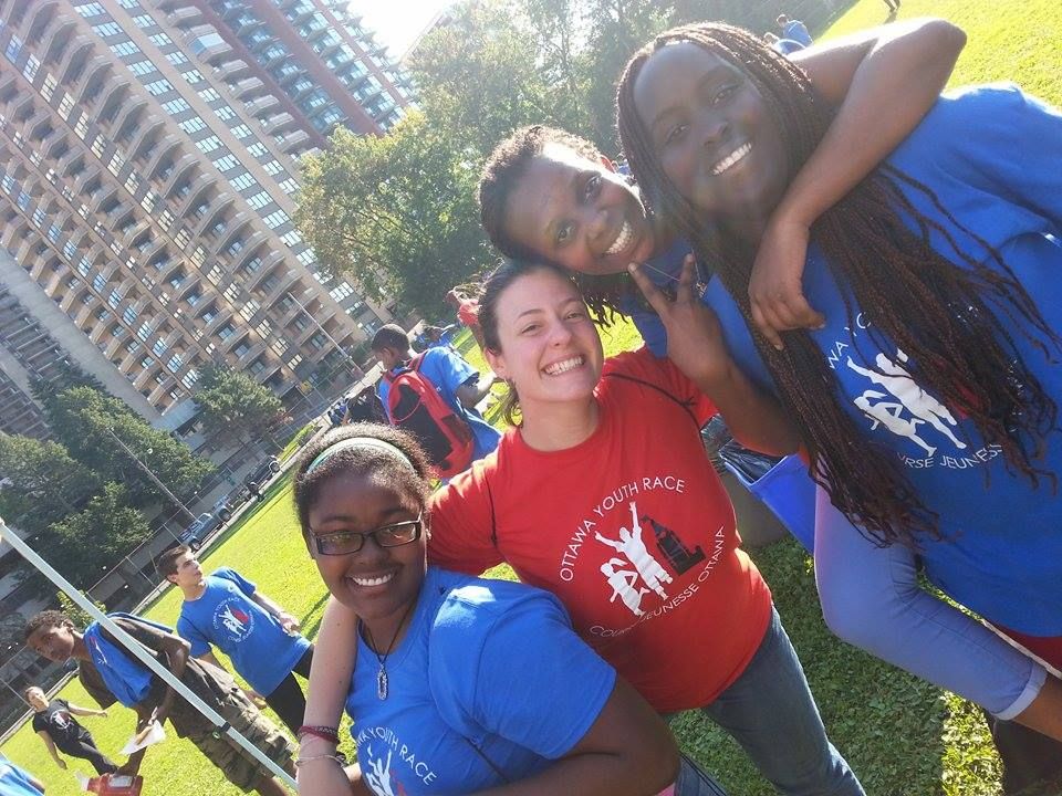 Oblique colour photograph of three black teenage girls in blue T-shirts, and a young adult white woman in red T-shirt with the logo and name of the Ottawa Youth Race event. In the background, a fenced-in field occupied by other young people and large residential buildings.