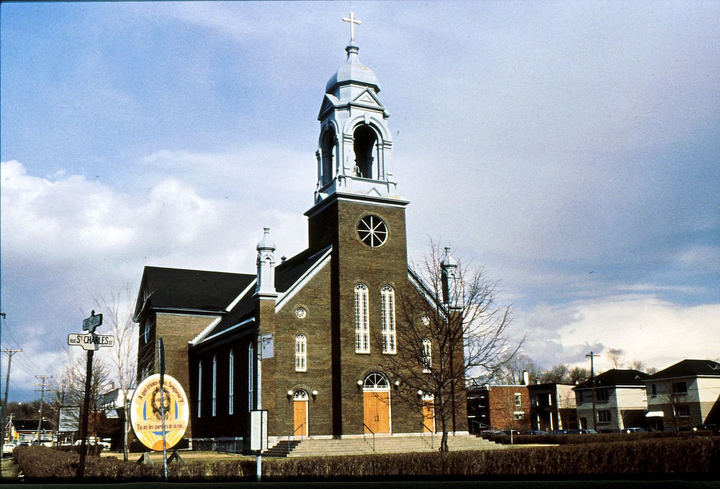 Colour photograph of a dark brown brick church with a striking silver bell tower surmounted by a cross. The facade of the church has three doors, four long windows, and a large bull’s-eye window. The church is located near a busy street, in a residential area.