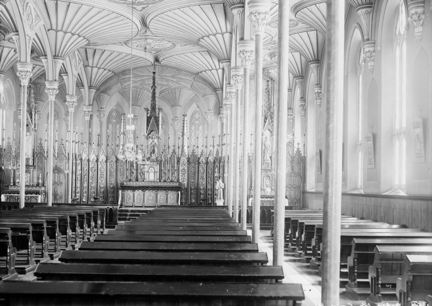Black and white photograph of the interior of a small church, with many benches in rows. The vaulted ceiling, made of carved wood, is supported by thin columns. The altar is very ornate.