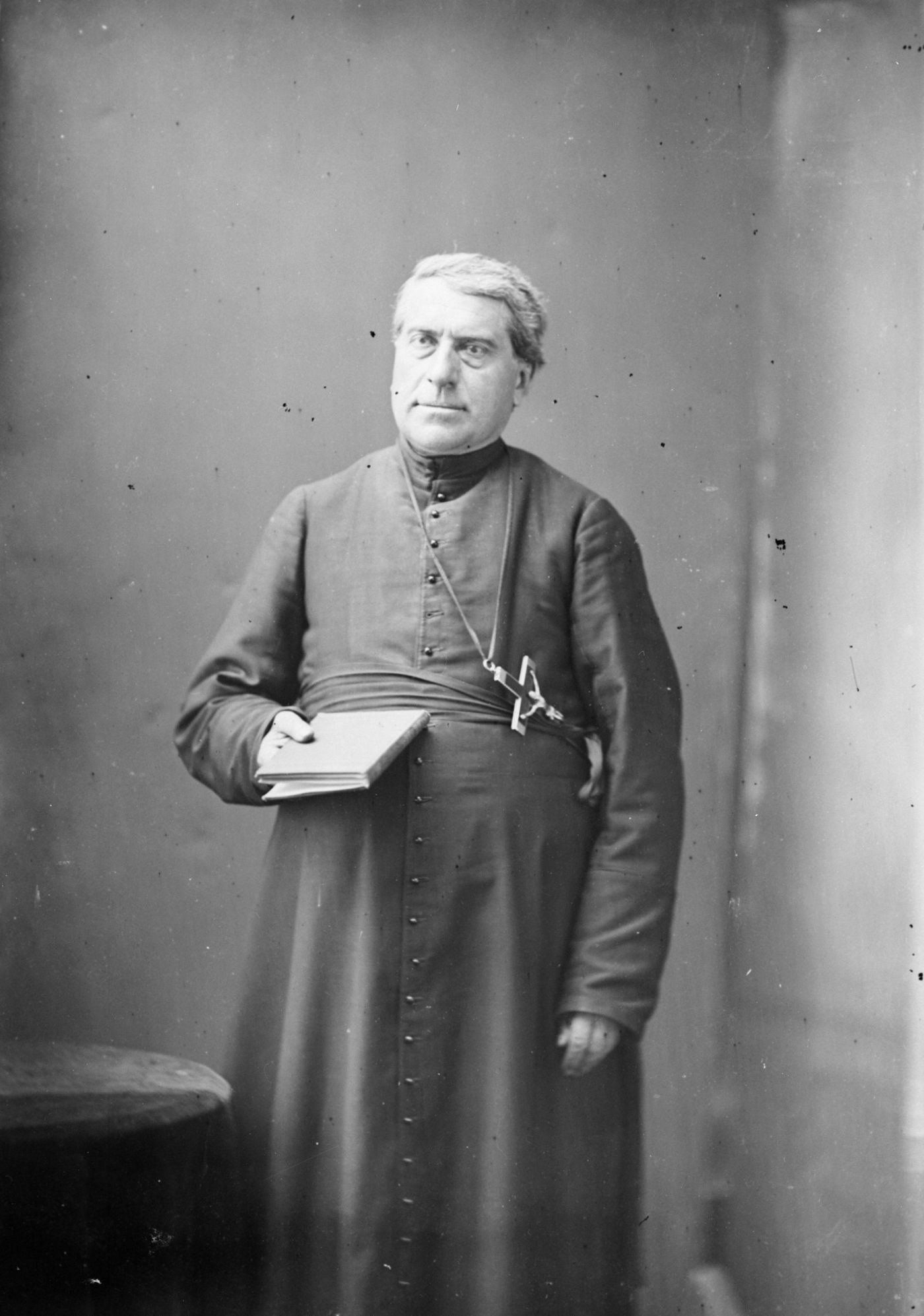 Black and white studio photograph of a mature priest wearing a cassock and a large cross. He is standing, holding a book in his right hand.