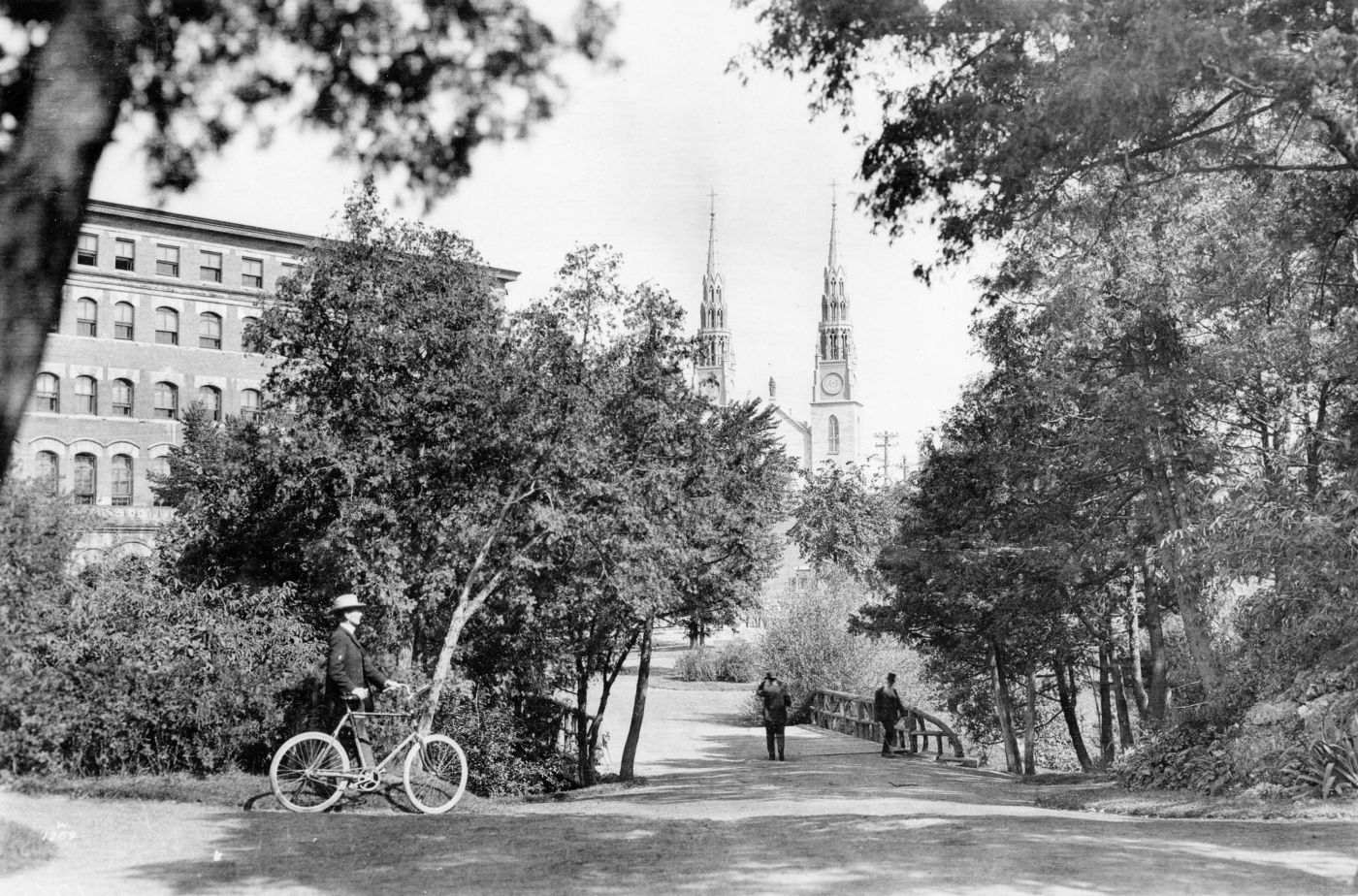 Black and white photograph showing a man walking beside his bicycle. He is moving toward a tree-lined esplanade leading to a wooden bridge. Two men are crossing the bridge on foot. Two steeples of a large church are visible in the background between the trees.