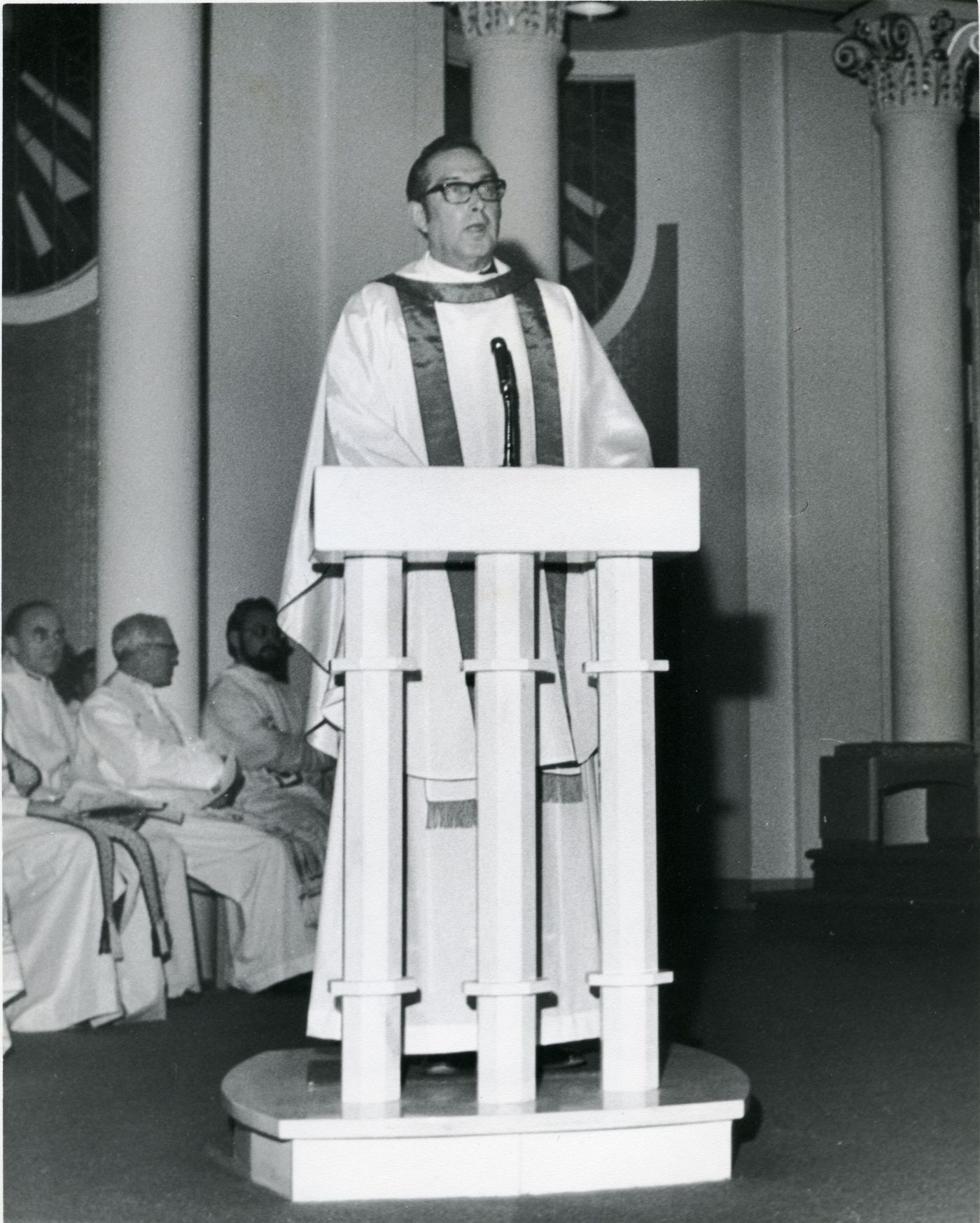 Photographie en noir et blanc d’un prêtre vêtu d’habits sacerdotaux blancs debout à la chaire d’une église.