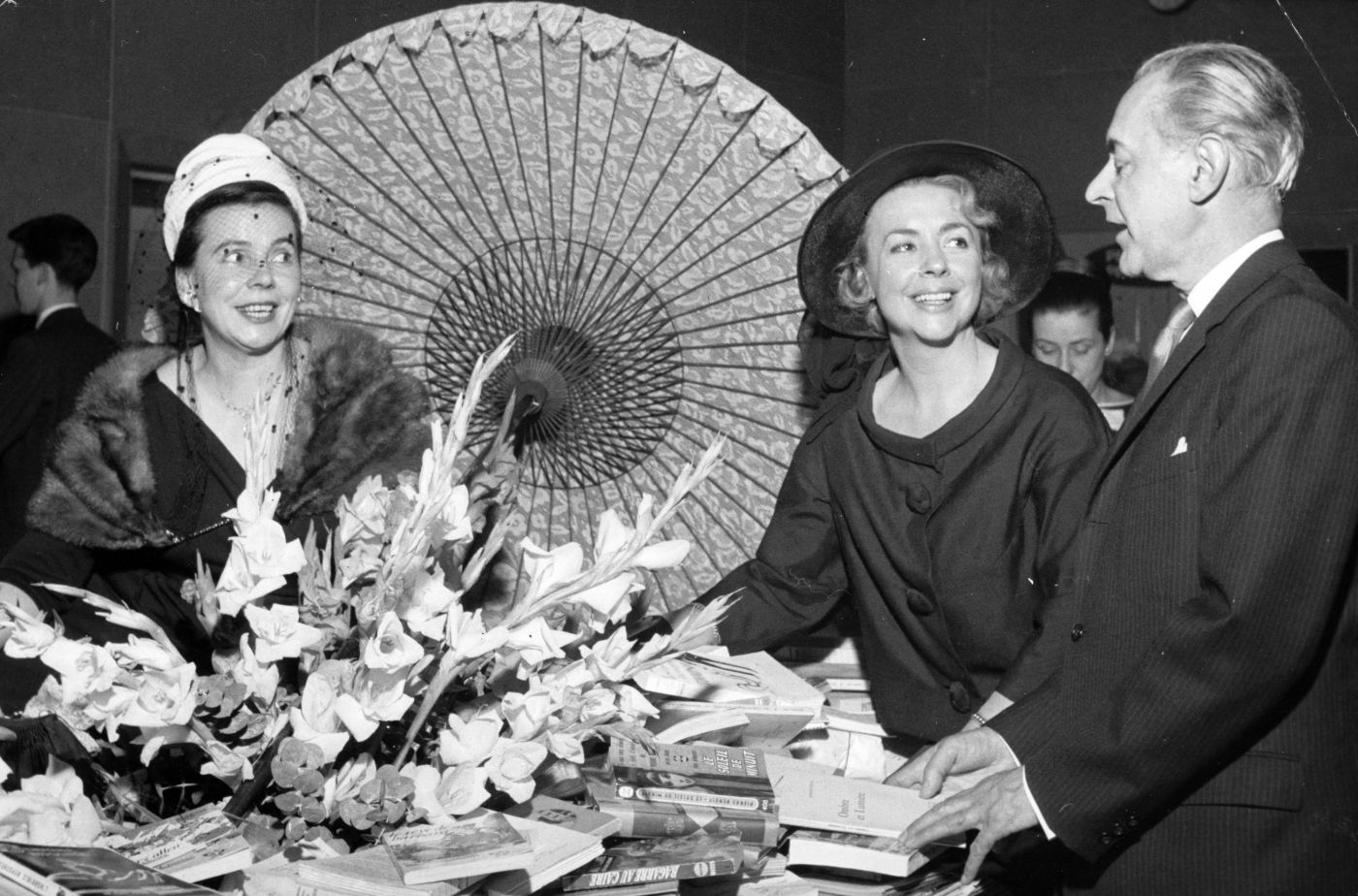 Photographie en noir et blanc de deux élégantes femmes d’âge moyen et d’un homme d’âge mûr, de chaque côté d’une table remplie de livres. Sur la table sont posés un grand bouquet de fleurs et un parasol ouvert.