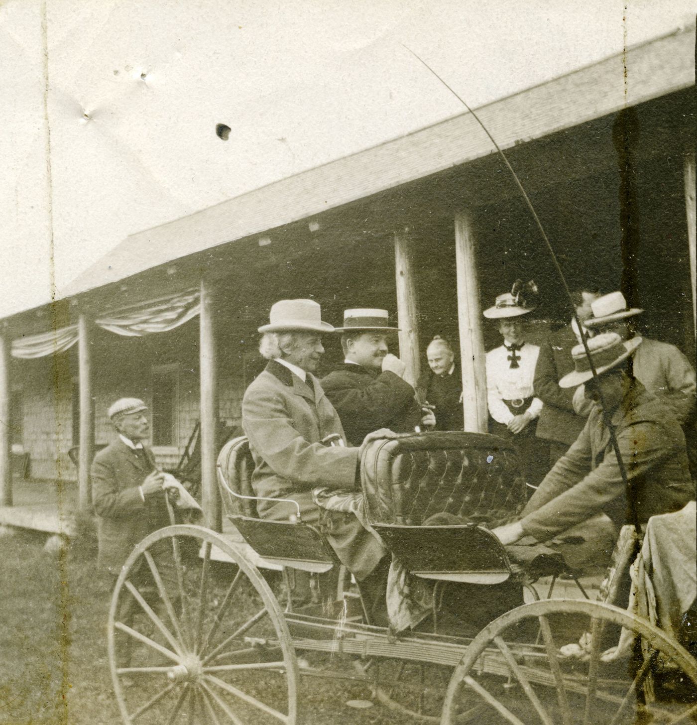 Black and white photograph of two well-dressed men sitting in a buggy in front of a wooden building. The white hair of the first is visible under his hat. The second, younger, wears a mustache. A third man prepares the buggy. Other people are watching them.