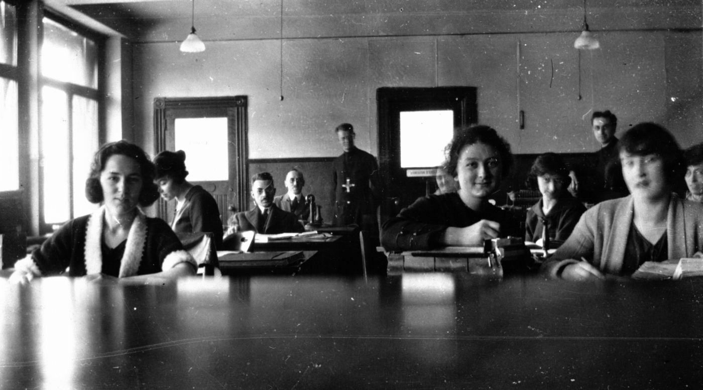 Black and white photograph of men and women sitting at desks. In the foreground, three young women. Standing at the back of the room, a man wearing religious garments.