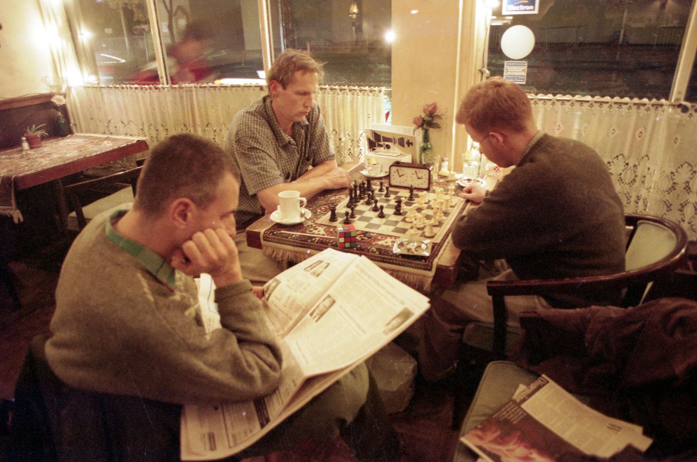 Photographie en couleur de trois hommes d’âge moyen, assis dans un café, le soir. Deux des hommes jouent aux échecs. Le troisième homme lit un journal à leur côté.