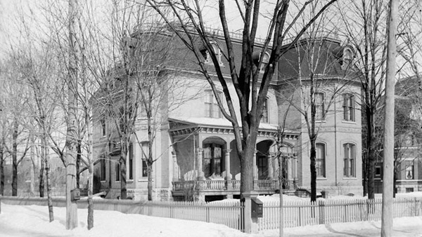 Black and white photograph of a three-story house with a front porch. The house, seen in winter, is set in a large yard surrounded by a fence. The yard and street are lined with trees.