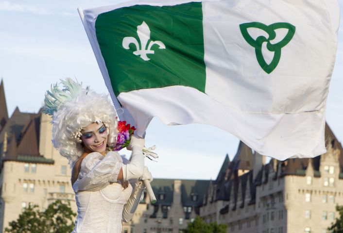 Colour photograph of a young woman, in three-quarter view, holding a wreath of flowers and a large Franco-Ontarian flag. She is dressed all in white and wears carnival attire. In the background, the Chateau Laurier, Ottawa's iconic hotel.