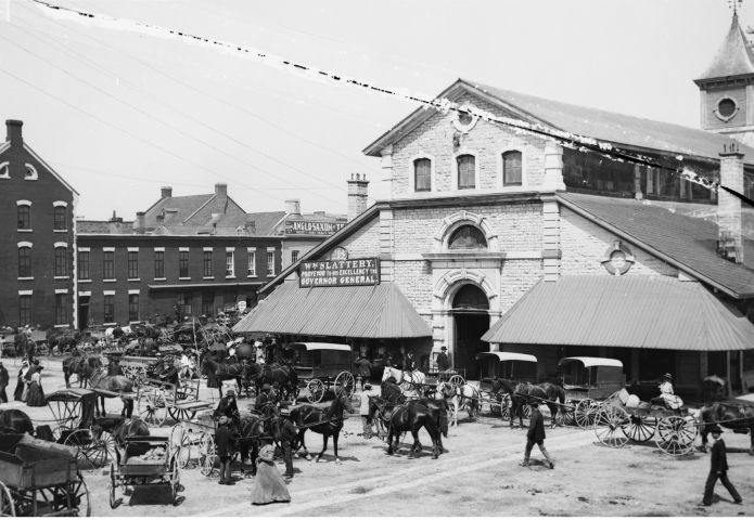 Black and white photograph of a three-storey stone building with awnings. Many people, along with different types of horse-drawn conveyances, wait in the square in front of the building.