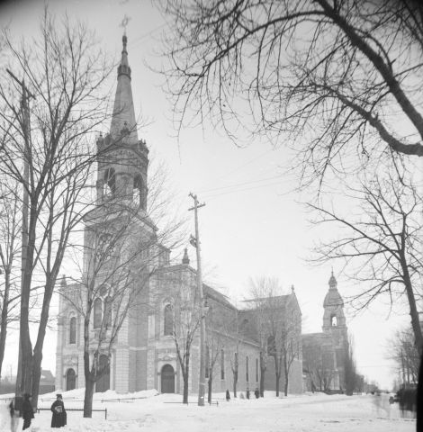 Photographie en noir et blanc d’une église vue en hiver. Une maison, des arbres et quatre personnes font partie du paysage. Une partie de la photographie est masquée, à cause de l’objectif photographique.