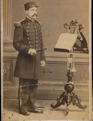 Sepia photograph of a middle-aged man wearing a uniform and a large mustache. He stands in front of a richly carved lectern which holds music scores and a trumpet. The name and title of the subject are handwritten at the bottom of the picture.