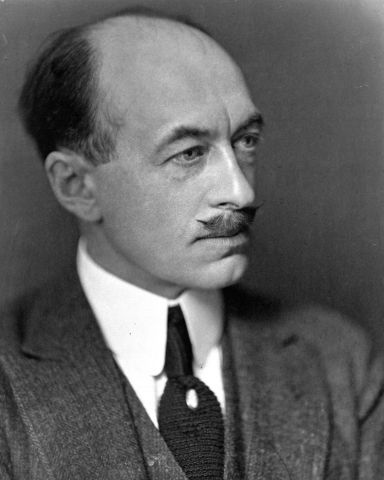 Black and white studio photograph of a mature, balding man with a mustache, in three-quarter view. He wears a gray suit, with a black tie and white tie pin, along with a serious expression.